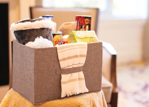 Holiday gift basket - mobile hero The holiday gift basket, filled with food and other home items sits on a chair in a room.