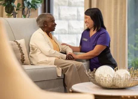 Amica Lounge Care A smiling Amica caregiver sitting beside an elderly woman on a lounge sofa, holding her hands warmly in a bright, comfortable common area