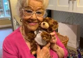 Amica Senior Resident Teddy Bear A cheerful elderly woman wearing glasses and a pink top smiles while holding a small teddy bear close to her