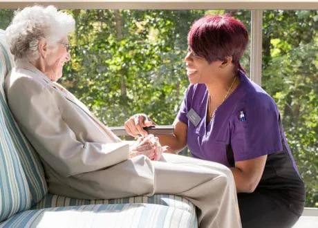 Amica Caregiver Senior Connection An Amica caregiver sits attentively beside an elderly woman near a window, holding her hands in a warm, engaged conversation