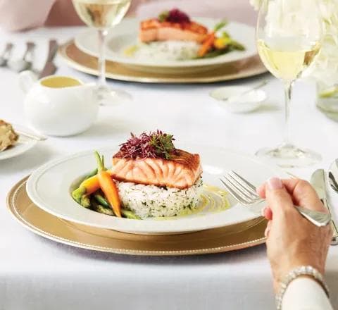Amica Dining - Salmon Plated Dinner An elegantly plated salmon fillet serving on a bed of rice with asparagus and roasted carrots, garnished with greens, on a plate with wine glasses and cutlery on a white linen table