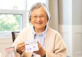 Amica Senior Arts Crafts Activity A smiling elderly woman wearing glasses holds up a small decorated card or artwork while seated near a bright window