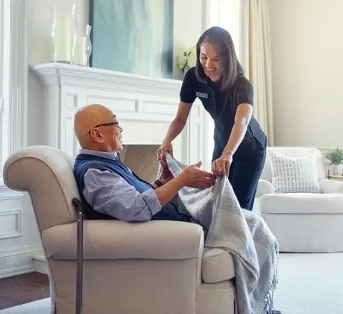 Amica Care Caregiver A smiling Amica caregiver draping a blanket over an elderly man seated comfortably in an armchair in a bright, cozy living room